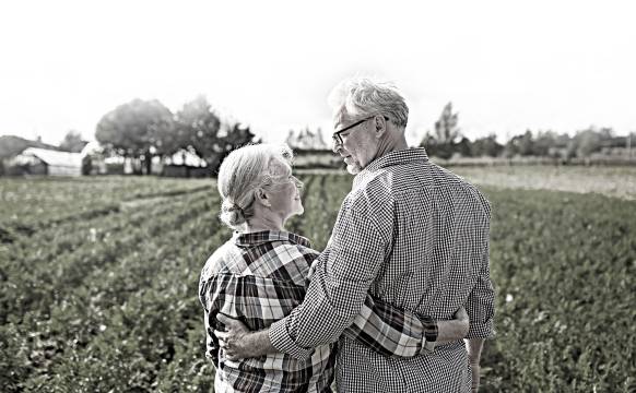 An elderly couple gaze into each others' eyes while standing in front of a field.