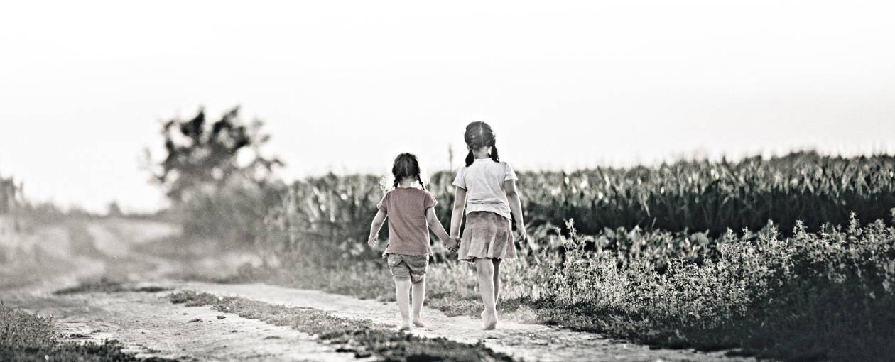 Two sisters walk down a dirt road near a corn field.