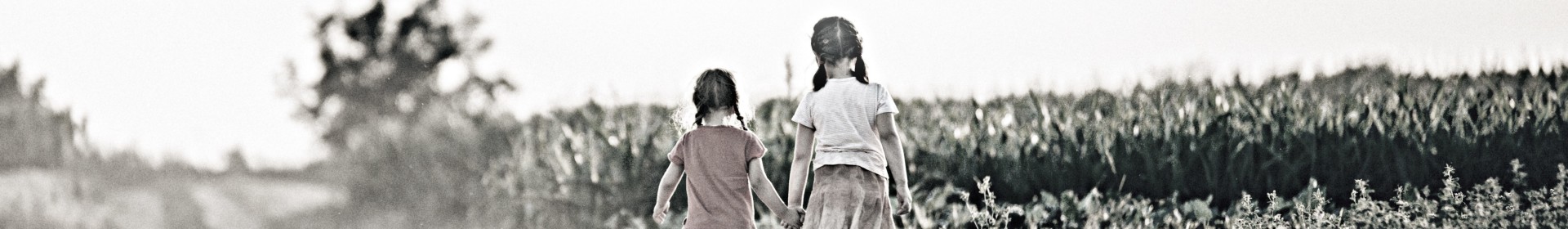 Two sisters walk down a dirt road near a corn field.